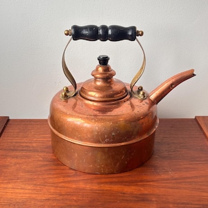 May include: A vintage copper tea kettle with a black wooden handle and brass accents. The kettle has a rounded body, a spout, and a lid with a black knob. It is sitting on a wooden surface.