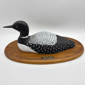 May include: A hand-carved wooden loon sculpture with black and white paint detailing. The sculpture is mounted on a circular wooden base with a brass plaque that reads "COMMON LOON GAVIA IMMER".