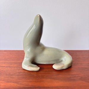 May include: A ceramic seal sculpture in a seated pose. The seal is a pale green colour with a glossy finish. The sculpture has a smooth, rounded form, with the head and body clearly defined. The seal's flippers are visible, and the sculpture is resting on a wooden surface.