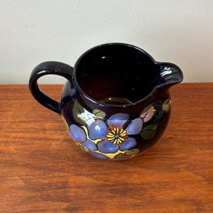 Op de afbeelding: Een donkerblauwe keramische kan met een handvat en tuit. De kan is versierd met blauwe, gele en paarse bloemmotieven. De kan staat op een houten oppervlak.