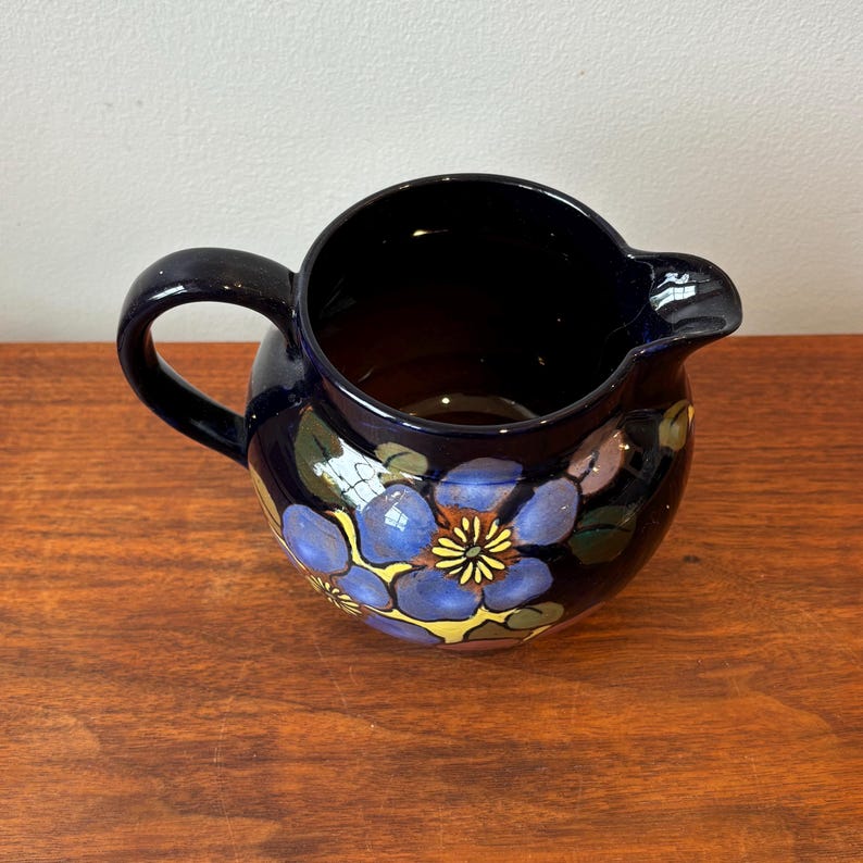 Op de afbeelding: Een donkerblauwe keramische kan met een handvat en tuit. De kan is versierd met blauwe, gele en paarse bloemmotieven. De kan staat op een houten oppervlak.