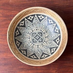 May include: A round ceramic bowl with a decorative design. The bowl features a central starburst pattern in black and cream, framed by a gold-coloured border. The rim has a textured, scalloped design. The bowl is set on a dark wooden surface.