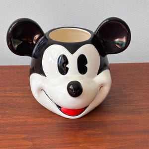 May include: A ceramic Mickey Mouse head-shaped planter. The planter features black ears, a black nose, and a red mouth. The face is white, and the interior is beige. A decorative item for Disney fans.