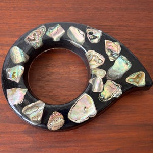 May include: A black, teardrop-shaped decorative object with a circular opening. The object is inlaid with iridescent abalone shell pieces. The shell pieces vary in size and shape, and are primarily white, green, and pink.