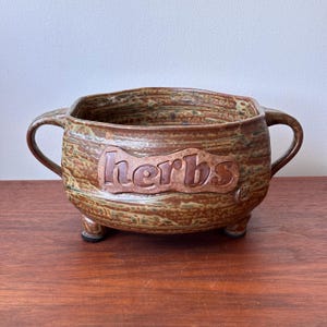 May include: A handmade ceramic herb pot with a textured brown and green glaze. The pot has two handles and three small feet. The word "herbs" is embossed on the front.