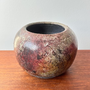 May include: A spherical ceramic bowl with a wide opening. The exterior has a mottled pattern of red, brown, and beige tones, giving it a rustic look. The interior is a dark, contrasting colour.