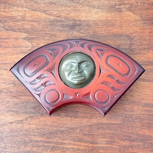 May include: A fan-shaped, carved wooden art piece with a central circular face. The face is a dark gray color, and the surrounding wood is a reddish-brown with intricate carved designs. The piece is displayed on a wooden surface.