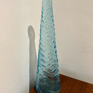 May include: A tall, light blue glass vase with a textured wave design. The vase is empty and has a rounded bottom.