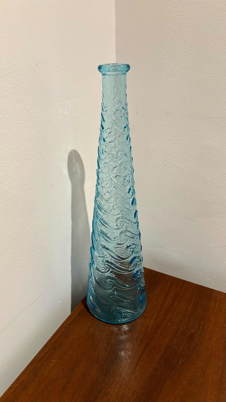 May include: A tall, light blue glass vase with a textured wave design. The vase is empty and has a rounded bottom.
