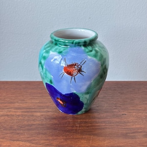 May include: A small, hand-painted ceramic vase with a round body and a narrow neck. The vase features a blue flower design with a red and orange center, set against a green background. The vase is sitting on a wooden surface.