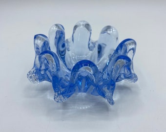 Puede incluir: Un pequeño cenicero de vidrio azul claro con un borde rizado. El cenicero está hecho de vidrio transparente con un tinte azul.
