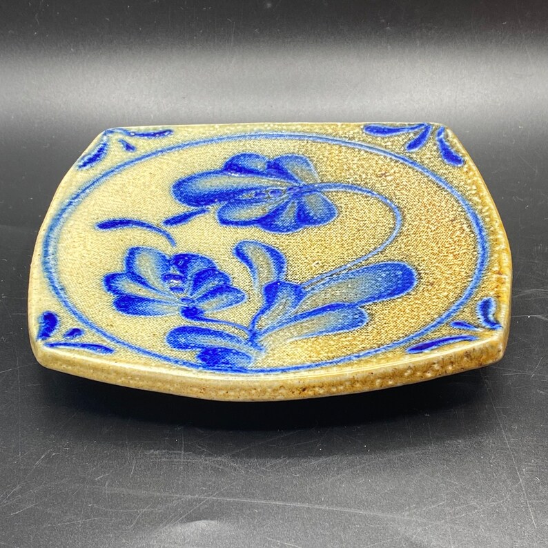K&ouml;nnte beinhalten: Ein quadratischer Keramikteller mit hellbrauner Glasur und blauem Blumendesign. Der Teller hat einen leicht erh&ouml;hten Rand und eine strukturierte Oberfl&auml;che.