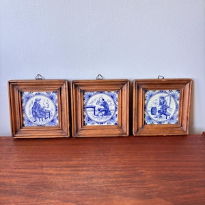 Può includere: Tre piastrelle in ceramica incorniciate con illustrazioni blu e bianche. Ogni piastrella presenta una scena diversa di persone in un ambiente tradizionale. Le piastrelle sono inserite in cornici di legno con un anello per appenderle.