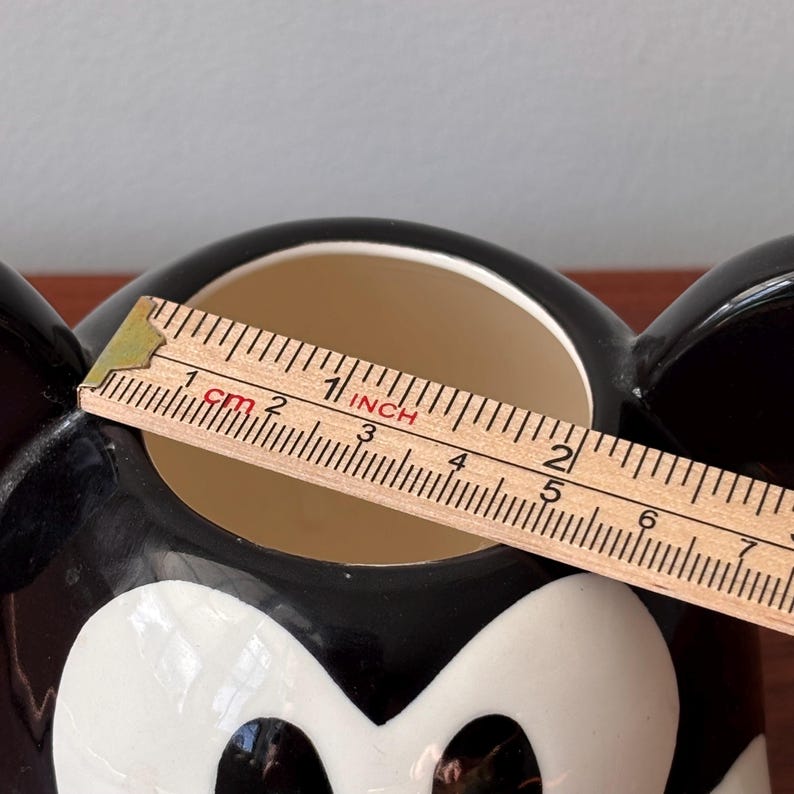 May include: A black and white ceramic mug shaped like Mickey Mouse's face. The mug has a beige interior and is being measured with a ruler. The ruler shows the mug's opening is approximately 2 inches (5 cm) wide.