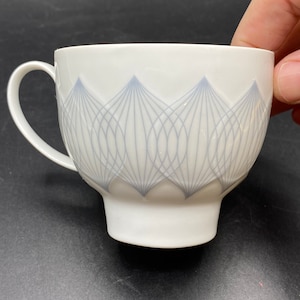 May include: A white ceramic teacup with a gray geometric design. The cup has a handle and a wide base.