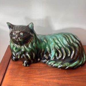 Könnte beinhalten: Keramik-Katzenfigur in liegender Pose. Die Katze hat eine glänzende Oberfläche mit einer Mischung aus dunkelbraunen und grünen Farbtönen. Das Fell ist mit strukturierten Pinselstrichen detailliert. Auf einer Holzoberfläche platziert.