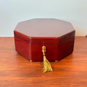 May include: A red, octagonal jewelry box with a glossy finish. The box has a gold-colored key and tassel. The box is sitting on a wooden surface. The box has small gold feet.