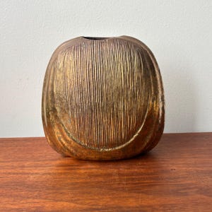 May include: A decorative, metallic vase with a textured, vertical-lined pattern. The vase has a rounded, oval shape and a dark opening at the top. The colour is a blend of gold, bronze, and hints of purple.