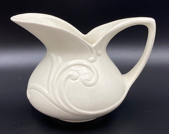 May include: A white ceramic pitcher with a decorative swirl design. The pitcher has a wide mouth and a handle.
