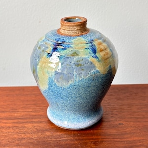 May include: A handmade ceramic vase with a rounded form and a narrow neck. The vase displays a mix of blue, green, and tan colours, creating a textured, glazed finish. The vase is placed on a wooden surface.