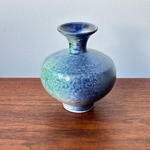 May include: A ceramic vase with a rounded body and a slender neck. The vase is predominantly blue with green and white highlights. The surface has a speckled texture, and the vase rests on a small, white base. The vase is a decorative item.