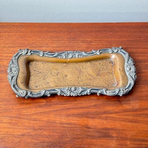 May include: A vintage rectangular tray with a silver-coloured ornate border and a gold-toned interior. The tray's interior features a subtle floral pattern. The border has detailed scrollwork and floral accents, adding to its vintage charm.