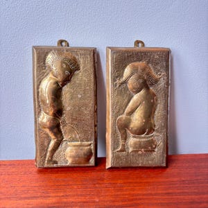 Peut inclure: Deux plaques en relief en laiton vintage. L'une représente un enfant urinant dans un pot, l'autre montre un enfant assis sur un pot de chambre. Les deux sont rectangulaires avec une boucle pour accrocher. Les plaques sont de couleur or antique chaud.