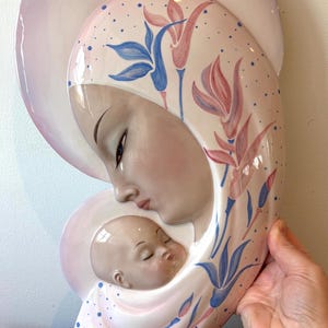 May include: A ceramic wall plaque depicting the Madonna and Child. The mother is holding the baby in her arms, and both have closed eyes. The background is a light pink with blue and pink floral designs.