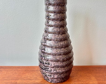 May include: A tall, textured vase with a dark purple and pink color scheme. The vase has a ridged design, with horizontal bands creating a layered effect. The vase is made of ceramic or a similar material.