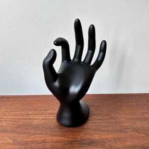 Può includere: Un espositore nero a forma di mano, con le dita estese in un gesto. Il supporto è realizzato in un materiale liscio e solido ed è progettato per contenere gioielli o altri piccoli oggetti. La mano è posizionata in una posizione rilassata e aperta.