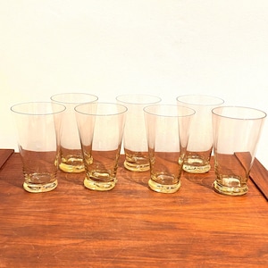 Pode incluir: Um conjunto de oito copos de bebida transparentes vintage com uma base amarelada. Os copos são altos, finos e ligeiramente alargados, colocados numa superfície de madeira.