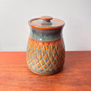 May include: A ceramic jar with a lid. The jar is orange with a diamond pattern and a dark teal band around the middle. The lid is orange with a small knob. The jar is sitting on a wooden surface.