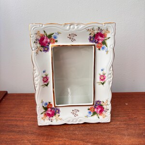 Peut inclure: Cadre photo en porcelaine blanche avec une ouverture rectangulaire. Le cadre est orné de motifs floraux colorés et d'une bordure dorée. Le cadre a un bord festonné.