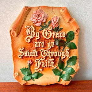 Op de afbeelding: Decoratieve plaat met de woorden "By Grace are ye Saved Through Faith" in sierlijke letters. De plaat heeft reliëf rozen en groene bladeren, tegen een perkamentachtige achtergrond met een warme, oranje tint.