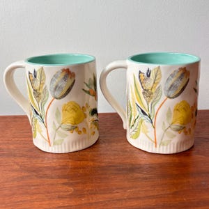 Puede incluir: Dos tazas de cer&aacute;mica con dise&ntilde;os florales. Las tazas tienen un exterior color crema con ilustraciones de flores amarillas, grises y naranjas. El interior de las tazas es de color verde azulado claro, y cada una tiene un asa curvada.