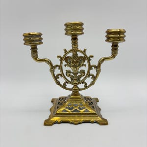 May include: A gold-toned brass candelabra with three candle holders. The candelabra features a decorative design with a face in the center and intricate scrollwork. The base is square with an openwork design.