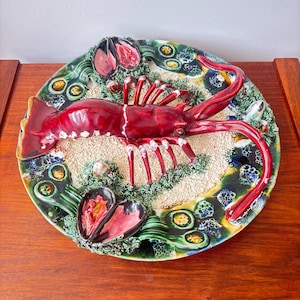 May include: A decorative ceramic plate featuring a vibrant red lobster as the centerpiece. The plate is adorned with green, blue, and yellow accents, along with two open mussels. The plate has a textured, sandy background.