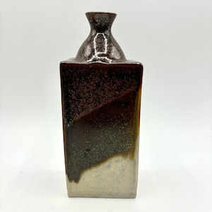 Puede incluir: Un jarrón de cerámica marrón y blanco con base cuadrada y cuello estrecho. El jarrón tiene un esmalte moteado con tonos marrones, verdes y blancos.