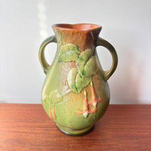 May include: A green ceramic vase with two handles and a brown rim. The vase features a raised floral design with green leaves and orange and white flowers. The vase is approximately 20 cm tall.