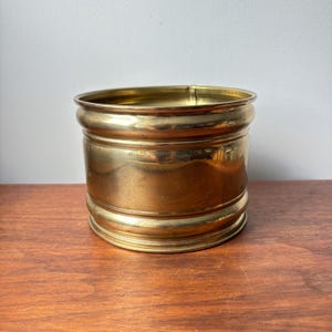 May include: A cylindrical brass planter featuring a polished metallic finish with a smooth, reflective surface. The vessel has a simple, unadorned design with a slightly flared rim and a matching base, showcasing a warm, golden-toned metal construction.