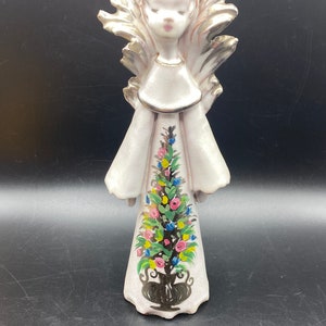 May include: A white ceramic angel figurine with gold wings and a painted Christmas tree on its dress. The tree is decorated with pink, blue, and yellow flowers.