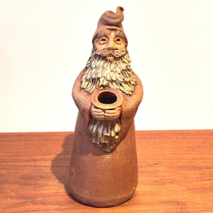 May include: A brown ceramic gnome figurine with a long beard and a pointed hat. The gnome is holding a small, round object. The figurine is standing on a wooden surface.