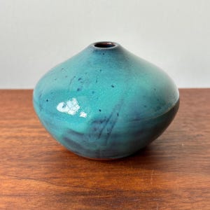 Op de afbeelding: Een kleine, turquoise keramische vaas met een afgeronde body en een smalle opening. De vaas heeft een glanzende afwerking met donkerblauwe en zwarte accenten en staat op een houten oppervlak. De vaas is een decoratief item.