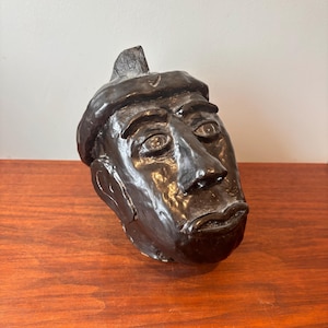May include: A dark brown sculpture of a human head wearing a beret. The sculpture has detailed facial features, including eyes, a nose, and a mouth. The head is tilted slightly, and the beret has a small, pointed detail on top.