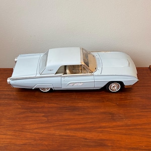 Puede incluir: Un coche modelo fundido a presión de color azul claro con techo blanco. El coche clásico tiene detalles cromados, neumáticos de banda blanca y el logotipo de Thunderbird. El modelo se exhibe sobre una superficie de madera.