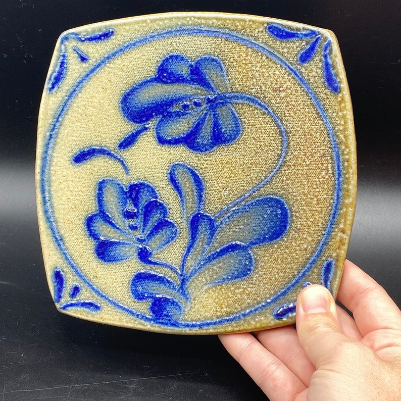 K&ouml;nnte beinhalten: Eine quadratische Keramikfliese mit cremefarbenem Hintergrund und blauem Blumendesign. Die Fliese zeigt zwei stilisierte Blumen mit Bl&auml;ttern und Stielen.