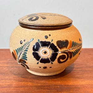 Puede incluir: Un tarro de cerámica redondo con tapa y acabado beige moteado. El tarro está decorado con diseños florales en blanco y negro, acentuados con detalles verdes y naranjas. La tapa tiene un borde marrón oscuro y un diseño floral a juego.