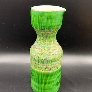 Puede incluir: Una jarra de cerámica verde con una superficie texturizada. La jarra tiene una boca ancha y un cuello estrecho.