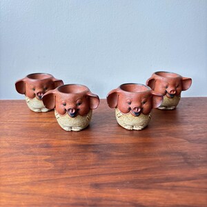 Może przedstawiać: Cztery ceramiczne doniczki w kształcie świni. Każda doniczka ma brązową świńską twarz z dużymi uszami, uśmiechniętym pyskiem i beżowym korpusem. Doniczki są ułożone na drewnianej powierzchni.