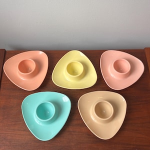 May include: A collection of five colorful egg cups in a mid-century modern style. Each cup is a different pastel color: peach, yellow, pink, teal, and beige. The egg cups have a triangular shape with a circular indentation for holding an egg.